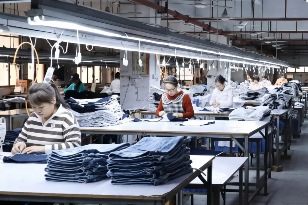 How to Verify a PFAS-Free Waterproof Fabric Supplier Before Placing an Order Garment factory operators assembling and processing sorted stacks of blue apparel at long tables on a bright industrial production line